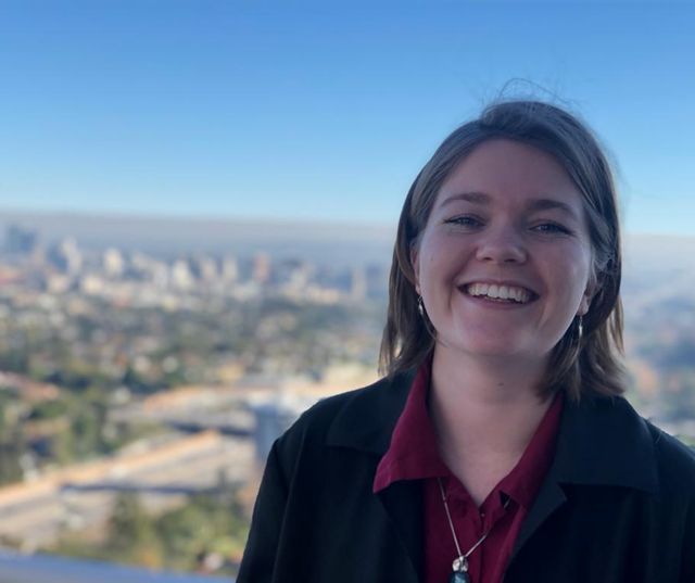 A picture of a smiling Rosie Tricks, who has dark hair a necklace and a red blouse, in front of a beautiful vista