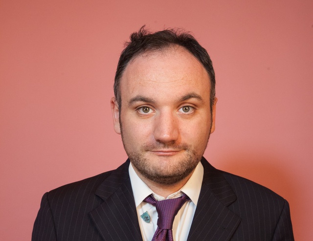 Headshot of James: A white man with dark hair and beard, against a pink background. He wears a suit a tie