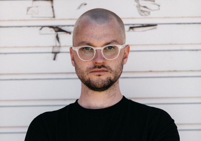 Head and shoulders shot of Adam against a white wall. He wears a black shirt, shaved head and white framed glasses
