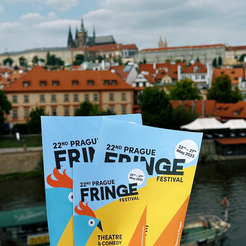 A hand holds up Prague Fringe flyers against the skyline of Prague
