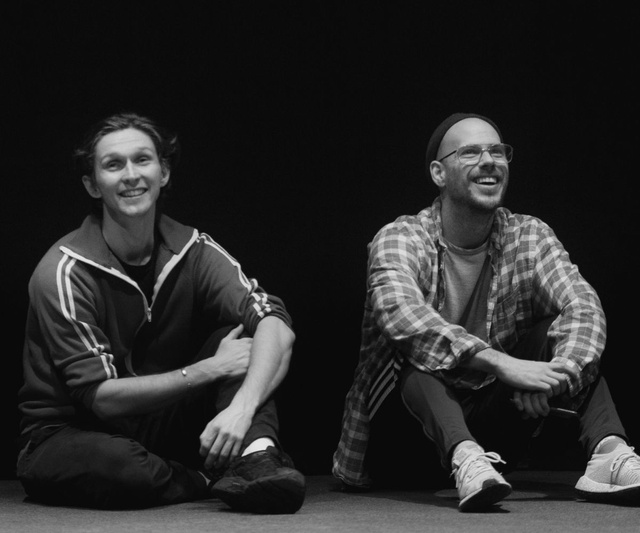 Black and White Photo - two men sit on the floor side by side smiling