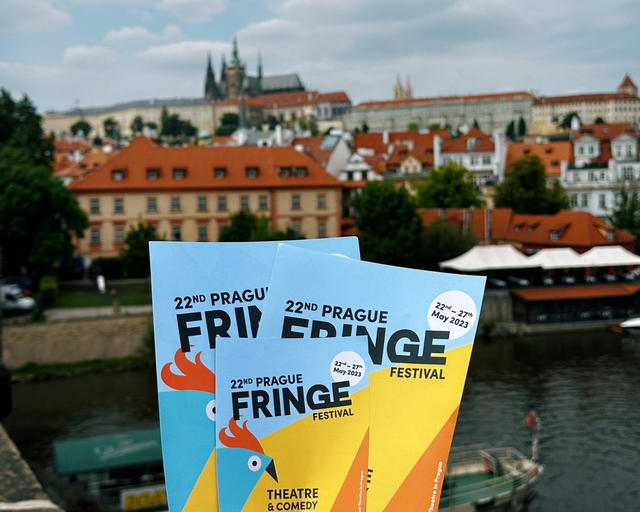 A hand holds up Prague Fringe flyers against the skyline of Prague