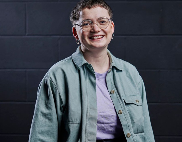 Ali stands in front of a black brick wall. Smiling and wearing glasses.