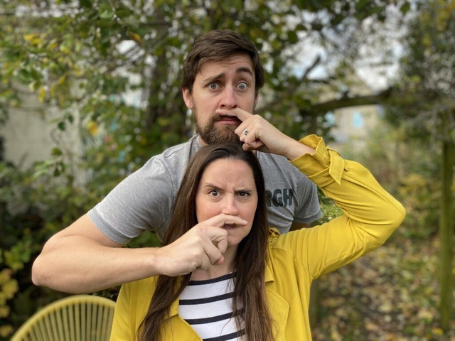 Heather stands in front of Feargus each making a moustache on the other's face with their fingers.