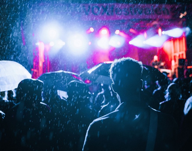 Silhouettes of people watching an outdoor stage at night, it's raining so umbrellas are up
