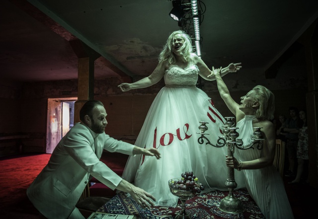 A theatre set with two women and a man stood around and on top of a dining table all dressed in white