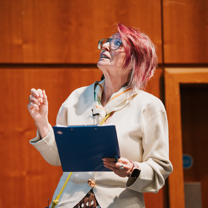 A white woman with red hair, glasses and a white top stands holding a laptop and presenting at a talk.