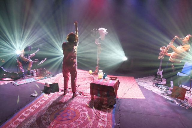 Taken from the back of the stage, floors are carpeted, stage lights beams are splayed out, an actor stands with their back to us in between a saxophonist and a guitarist
